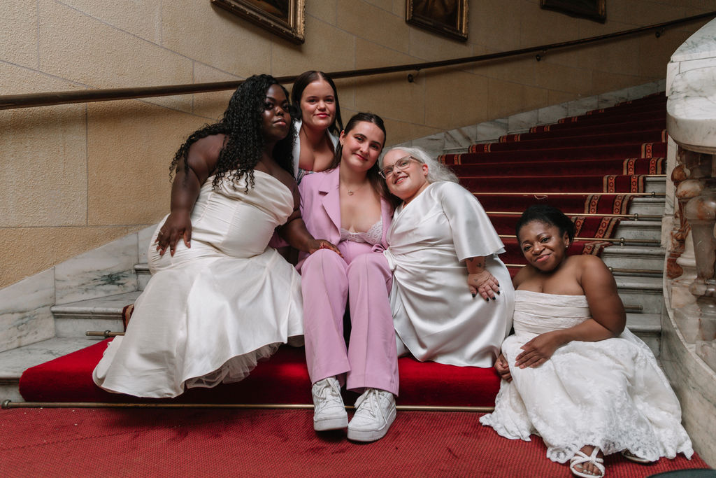 Chamiah is sat on a red staircase with 4 short stature individuals. The models had just walked London fashion Week, wearing Chamiah's bridal designs<br />
