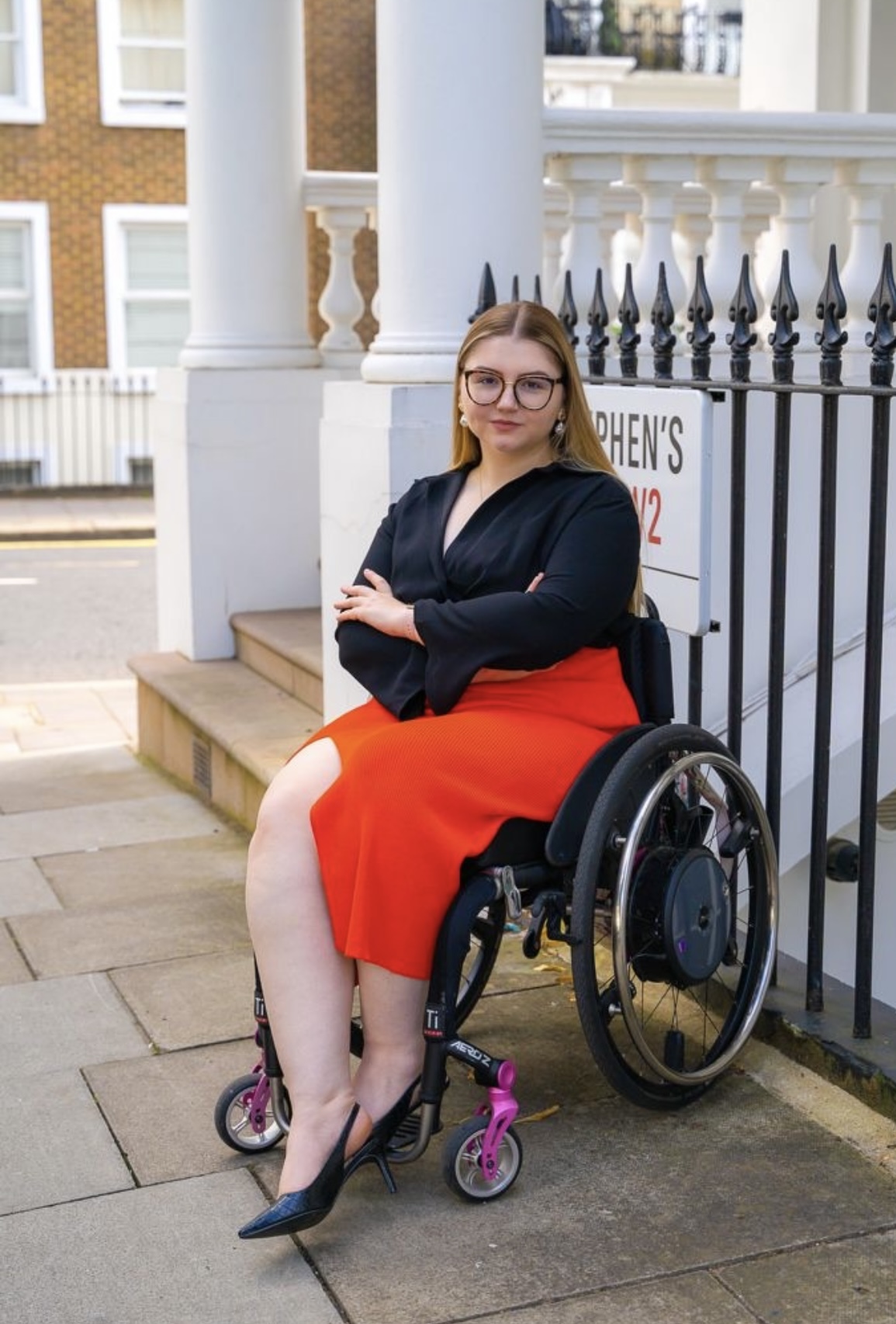 Charley is a 26 year old white woman. She is sitting in her tilite wheelchair, with emotion wheels, and the image is a full body image, she has her arms crossed and is staring at the camera. She has blonde highlighted hair, and brown eyes. She is wearing large framed glasses, and is wearing a black v neck shirt, with a red pencil skirt, with a split. Charley is also wearing black sling back heels. The background of the image shows black iron bars, and a london street sign.