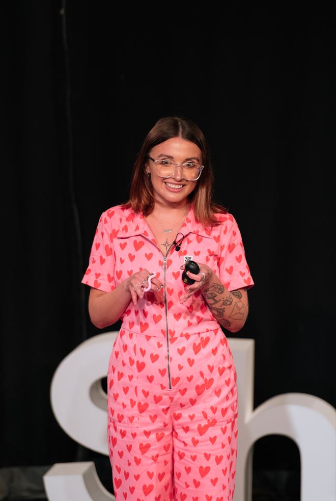 Ellie is a White woman with dark hair. She is speaking on stage wearing a pink jumpsuit with red hearts and holding a pink fidget toy.