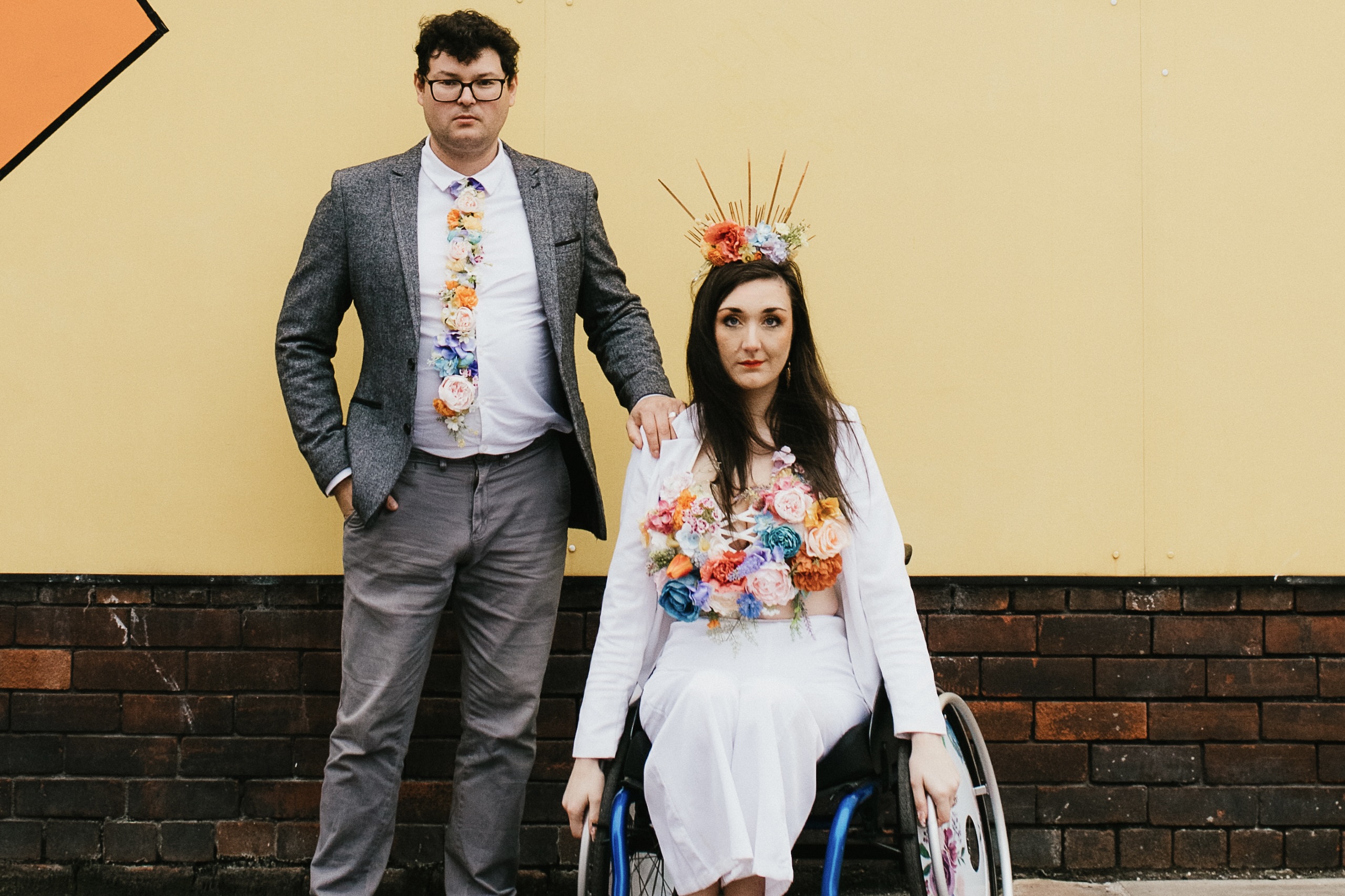 Georgina is a white, brunette female sat in a blue manual wheelchair. She looks intensely at the camera alongside her husband, Richard, who is a white brunette male. They wear colourful faux florals.<br />
