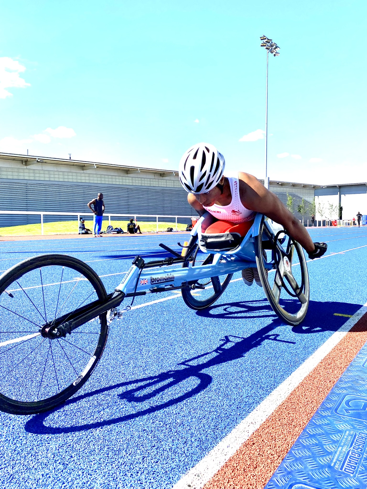 Black female pushing a sky blue racing wheelchair, wearing a white helmet and red and white Team England kit. Background- dark blue athletics track.