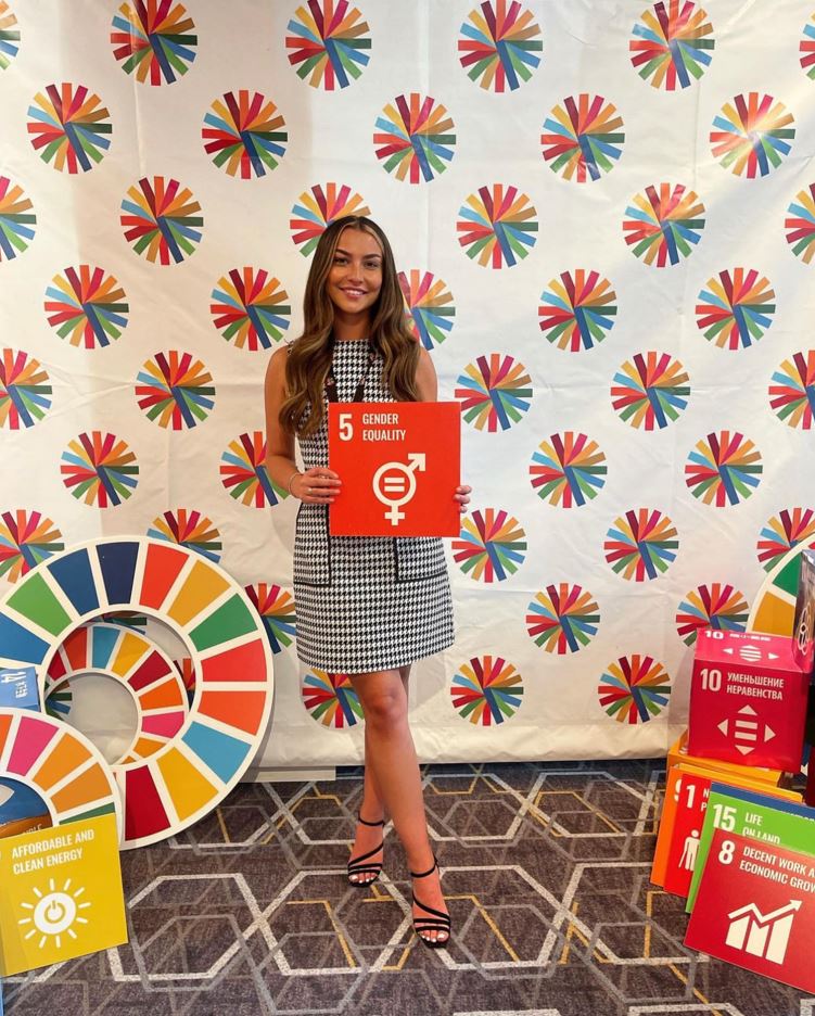 Kelly is a white woman with long brown hair and scattered blonde highlights. She is seen at the UN General Assembly Camp 2030 youth summit in New York holding an SDG 5: Gender Equality card. She is wearing a black checked dress and black high heeled sandals.<br />
