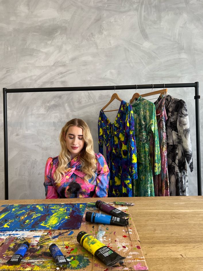 Kimberley wears one of her Warehouse dresses from the collection, a pink abstract patterned dress, and sits at a table with acrylic paints.<br />
