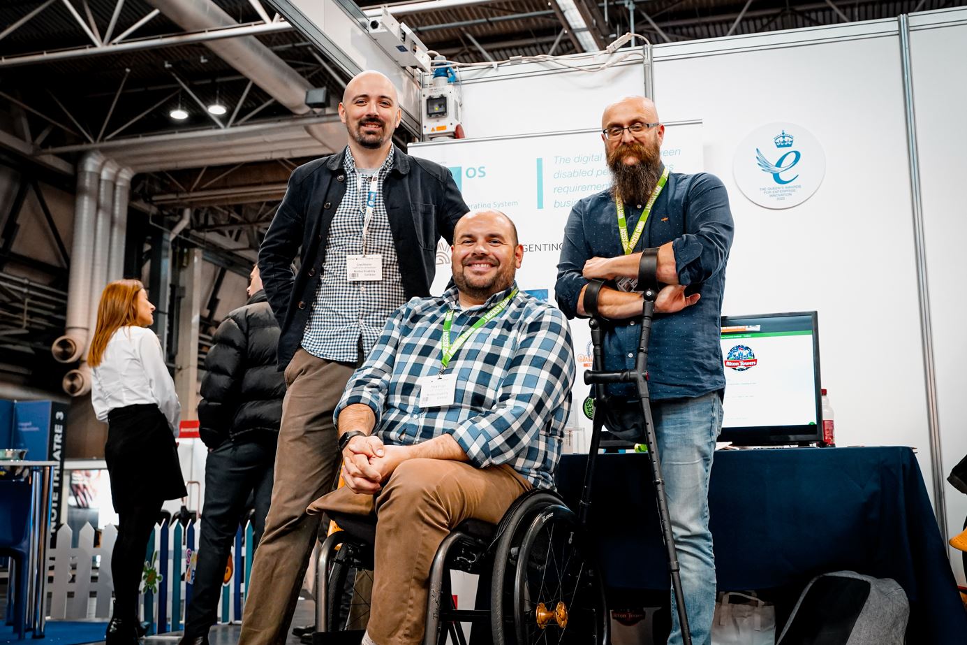 Mark with colleagues Martin and Greg at a conference stand. All 3 are white men with little hair and beards, dressed casually and smiling. Mark is a wheelchair user, whilst Martin is using crutches.