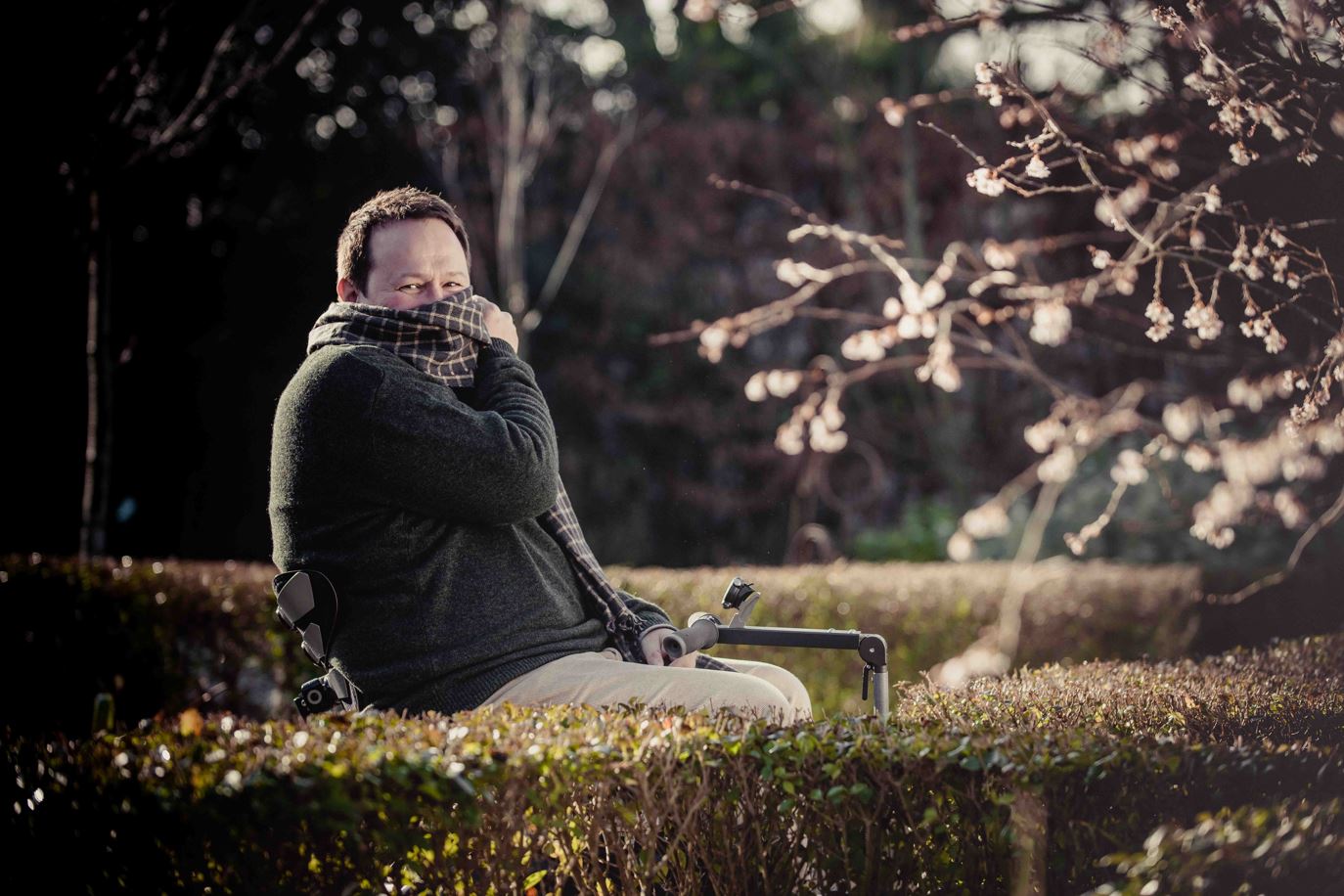 Mark Lane in an autumn garden, wearing a dark green jumper and a woollen brown scarf. Mark leans back in his chair and flicks the scarf around the lower part of his face, in a cheeky style.