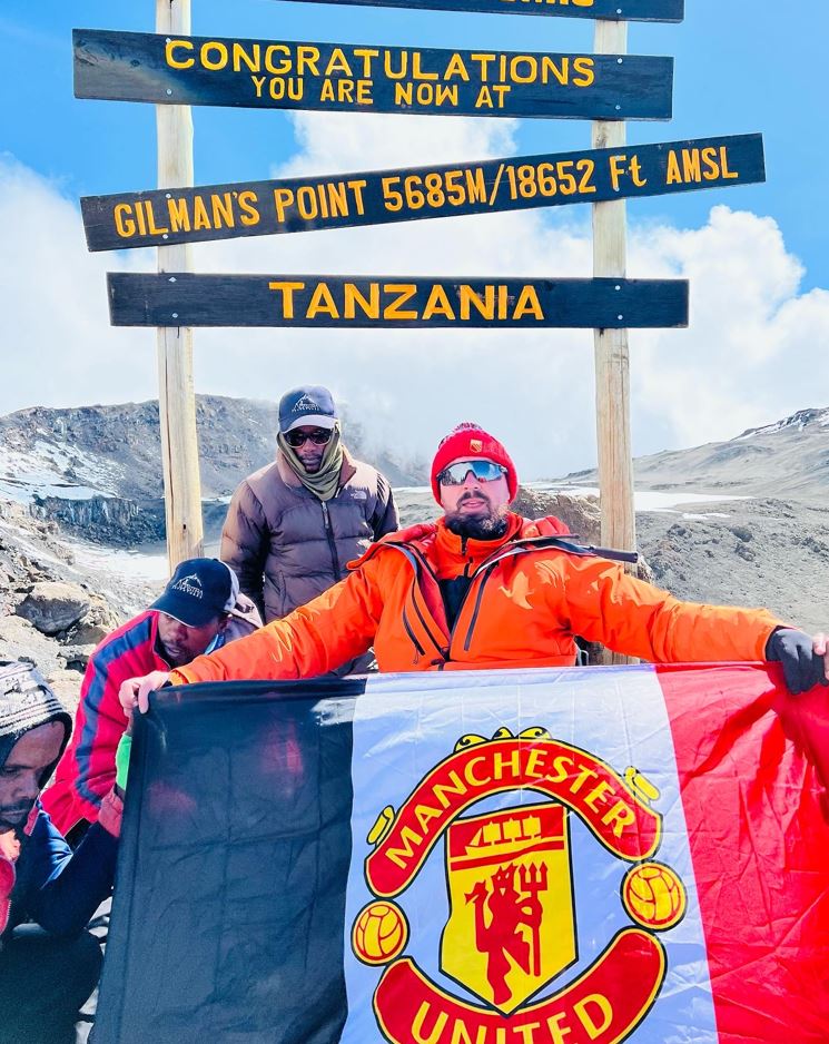 Bearded man with bobble hat and sunglasses dressed in Mountain Summit clothing and sat in his adapted Mountain Trike posing with his beloved Manchester United flag at the summit of Mount Kilimanjaro in June 2022<br />

