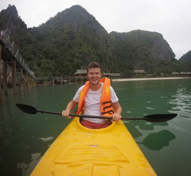 Angus is wearing an orange life jacket and is sat in a yellow canoe holding an oar on a river with mountains in the background.