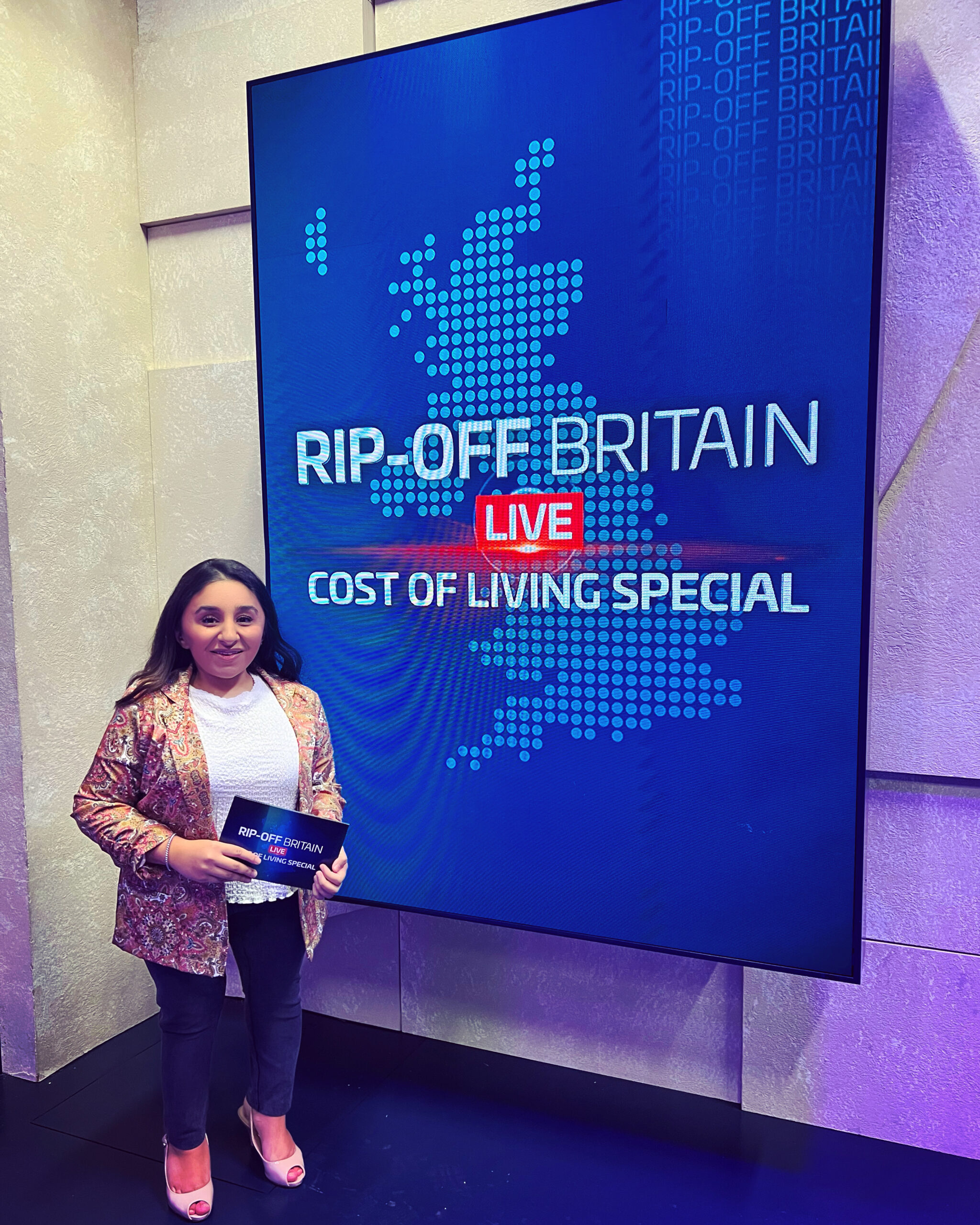 Shani Dhanda stands in front of a large blue digital screen with image of the UK.  It says 'Rip-Off Britain Live, Cost of Living Special', the same as the card Shani is holding. Shani is a South Asian short-stature woman in her mid-thirties with long brown hair.  She wears a gold brocade jacket, white top, blue jeans and nude slingback shoes.  